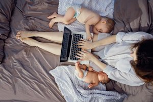 Frau sitzt mit zwei Babys im Bett und schreibt auf ihrem Laptop